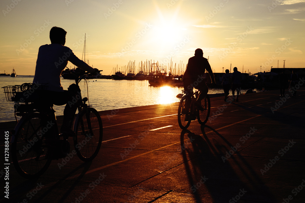 Beautiful quay near the sea on sunset. Silhouettes of people riding bicycles near the water. Ships, yachts and catters moored to the pier.
