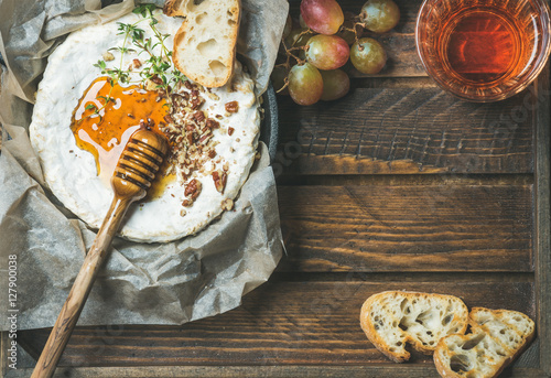 Homemade camembert cheese with honey, herbs and nuts, green grapes, baguette slices and glass of rose wine over rustic wooden background, top view, copy space