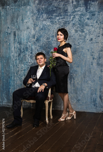 a man in a blue suit with a wine glass sits on a chair brunette woman in a black dress with a rose standing next