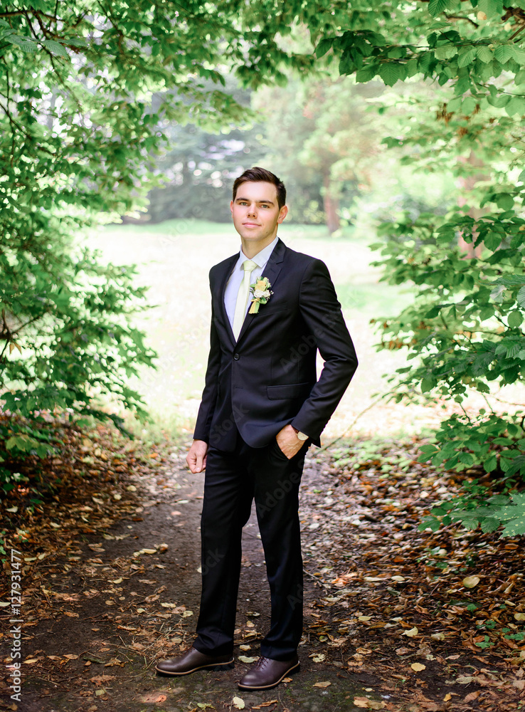 young handsome groom in a black suit standing alone outdoors