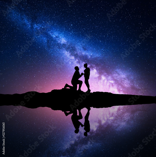 Milky Way. Silhouettes of a man making marriage proposal to his girlfriend on the hill near the lake with starry sky reflection in water. Night landscape. Silhouette of couple. Milky way with people