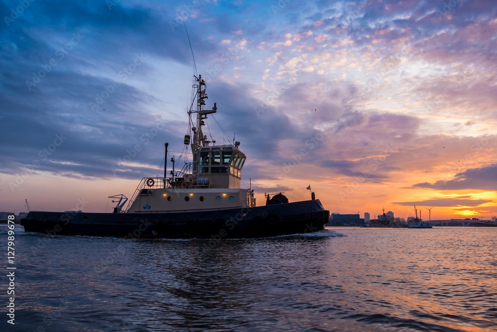 Tugboat is sailing during a beautiful sunset in port. Colorful clouds at the background.
