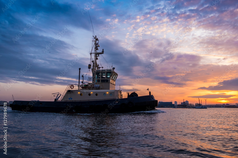 Fototapeta premium Tugboat is sailing during a beautiful sunset in port. Colorful clouds at the background.
