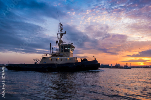 Tugboat is sailing during a beautiful sunset in port. Colorful clouds at the background.