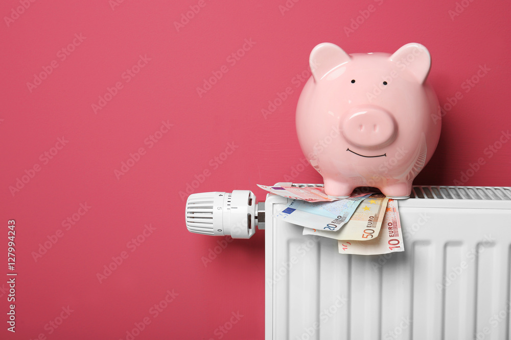 Obraz premium Savings concept. Piggy bank and money on heating radiator with temperature regulator on pink background