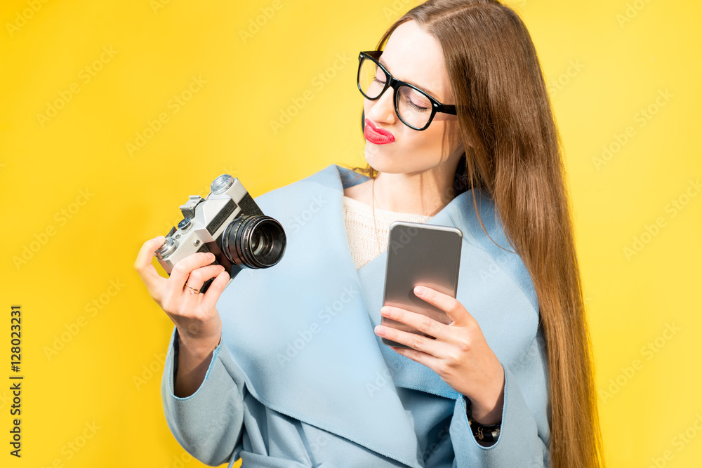 Confused nerdy woman photographer with retro camera and phone on the yellow wall background Confused nerdy woman photographer with retro camera and phone on the yellow wall background