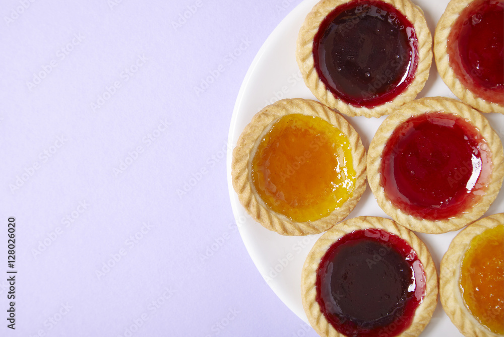 Overhead view of a plate full of assorted fruit jam tarts on a pastel purple background with blank space at side Overhead view of a plate full of assorted fruit jam tarts on a pastel purple background with blank space at side