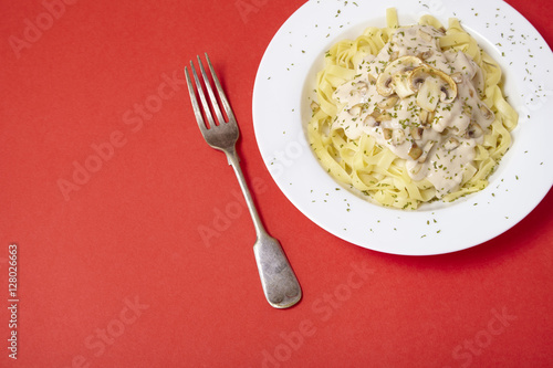 A dinner dish full of tagliatelle spaghetti with a creamy mushroom pasta sauce, on a bright red background with fork and blank space at side