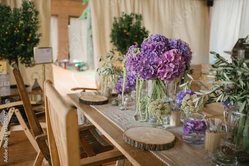 White and violet peonies stand in glass bottles on wooden dinner