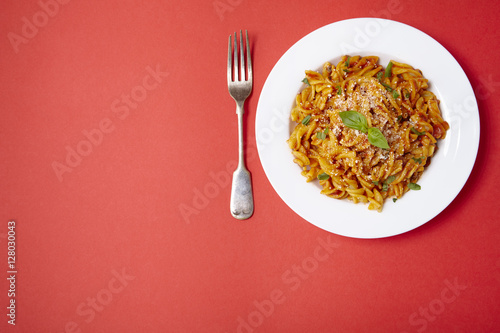 A dinner dish full of tomato and basil fusilli pasta on a bright red background with fork and empty space at side
