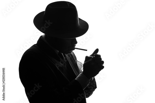 Silhouette of private detective lights cigarette. Agent looks like Al Capone stay side to camera. Police criminal scene in black and white. Gangster studio shot