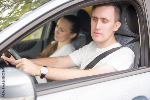 Young male driver looking at the side view wing mirror to see areas behind, woman passenger near
