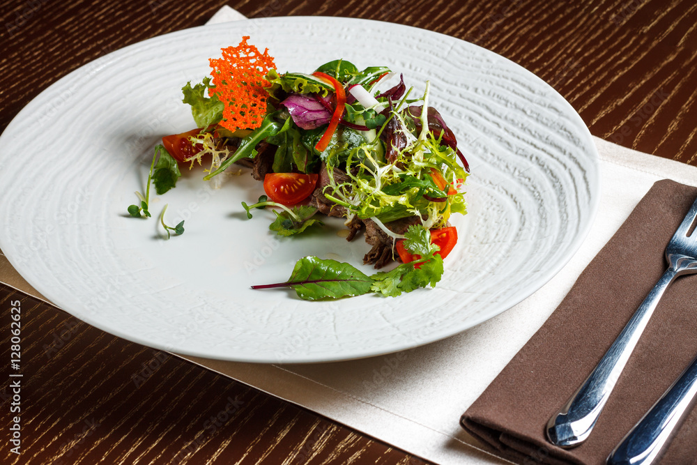 Vegetable salad menu for the restaurant in a large white plate on a white table cloth on a wooden table. Salad on the table with knife and fork. Svezhive vegetables. Vegetable salad menu for the restaurant in a large white plate on a white table cloth on a wooden table. Salad on the table with knife and fork. Svezhive vegetables.