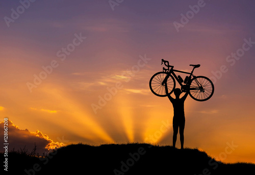 Silhouette the man stand in action lifting bicycle above his head on the meadow on sunset