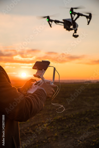 Man controls the fly of quadrocopter in field over sunset background