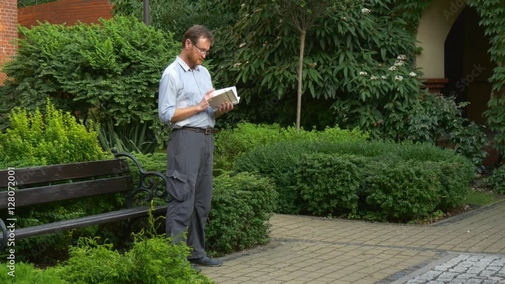 Elderly Man Stands Near to a Small Wooden Bench and Holds Book by a Hands Elderly Man Stands Near to a Small Wooden Bench and Holds Book by a Hands