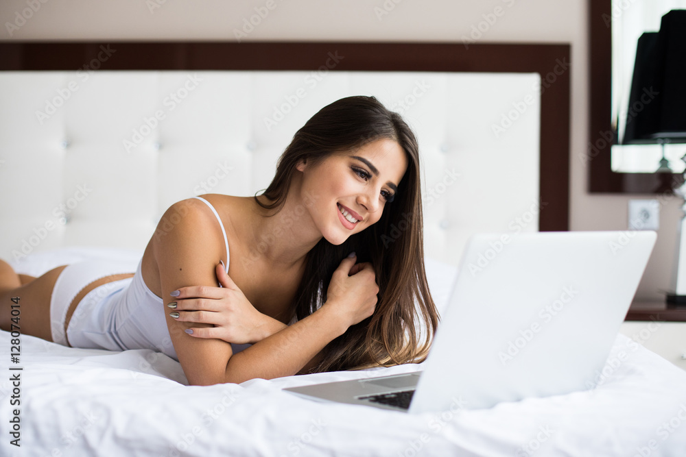 Smiling woman catching up on her social media as she relaxes in bed with a laptop computer on a lazy day Smiling woman catching up on her social media as she relaxes in bed with a laptop computer on a lazy day