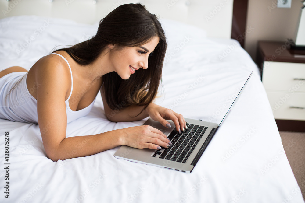 Smiling woman catching up on her social media as she relaxes in bed with a laptop computer on a lazy day Smiling woman catching up on her social media as she relaxes in bed with a laptop computer on a lazy day