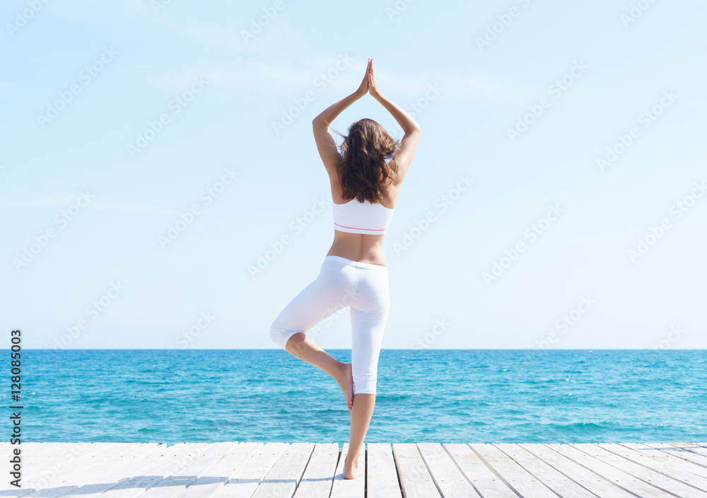 Woman in white sporty clothes meditating on a wooden pier on summer.  Sea and sky background. Yoga, sport, vacation, traveling and freedom concept.