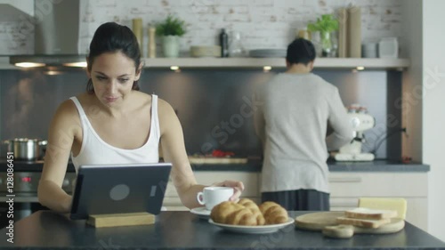 Beautiful Young Woman Sits at the Table in the Kitchen while Uses Tablet Computer, Sips Coffee and Eats Croissants. Young Man in the Background Cooks. Shot on RED Cinema Camera in 4K (UHD). photo