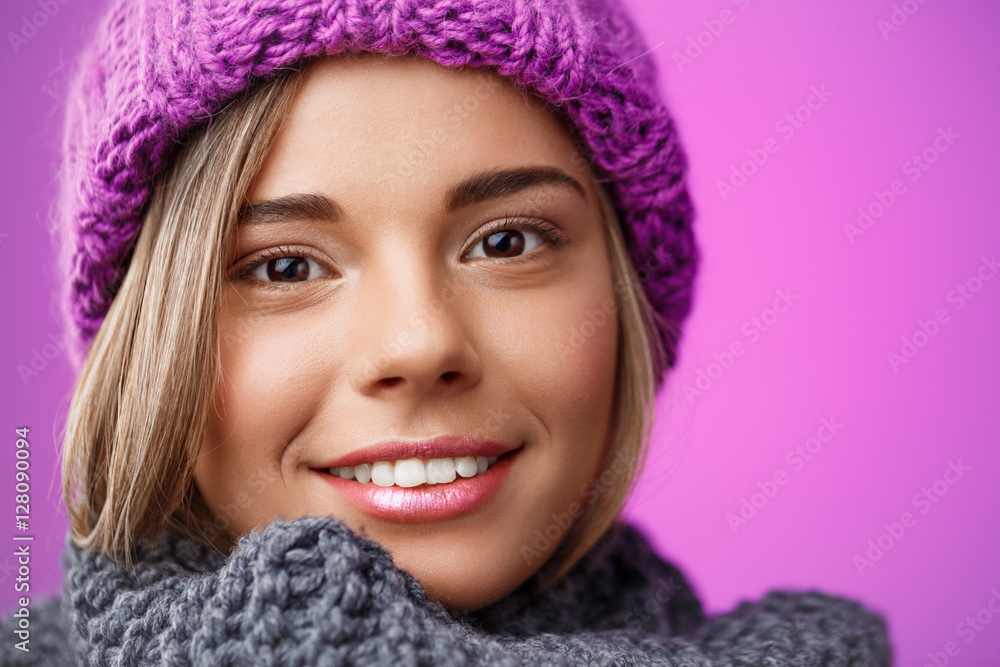 Young beautiful fair-haired girl in knited hat and sweater smiling looking at camera over violet background. Young beautiful fair-haired girl in knited hat and sweater smiling looking at camera over violet background.