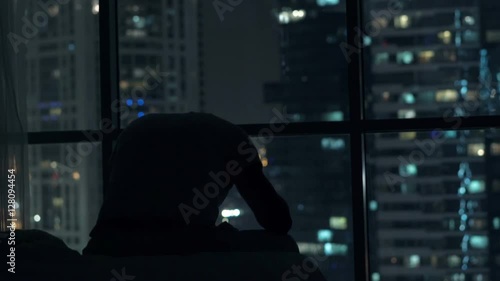 Sad, unhappy man sitting on bed close to the window during night 
