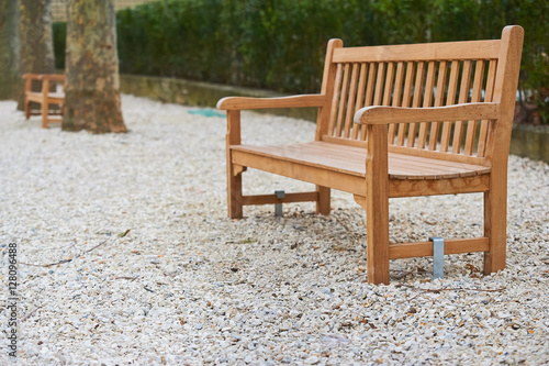 Wooden light brown bench at park surrounded by pebbles, trees, greenery © sebastiangora