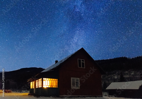 Wooden house in winter forest with lightning window in night at the stars night sky background. Christmas or New Year night scene.
