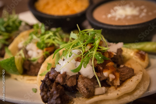 Plate of three street style tacos topped with micro greens, onions, chives and various sides