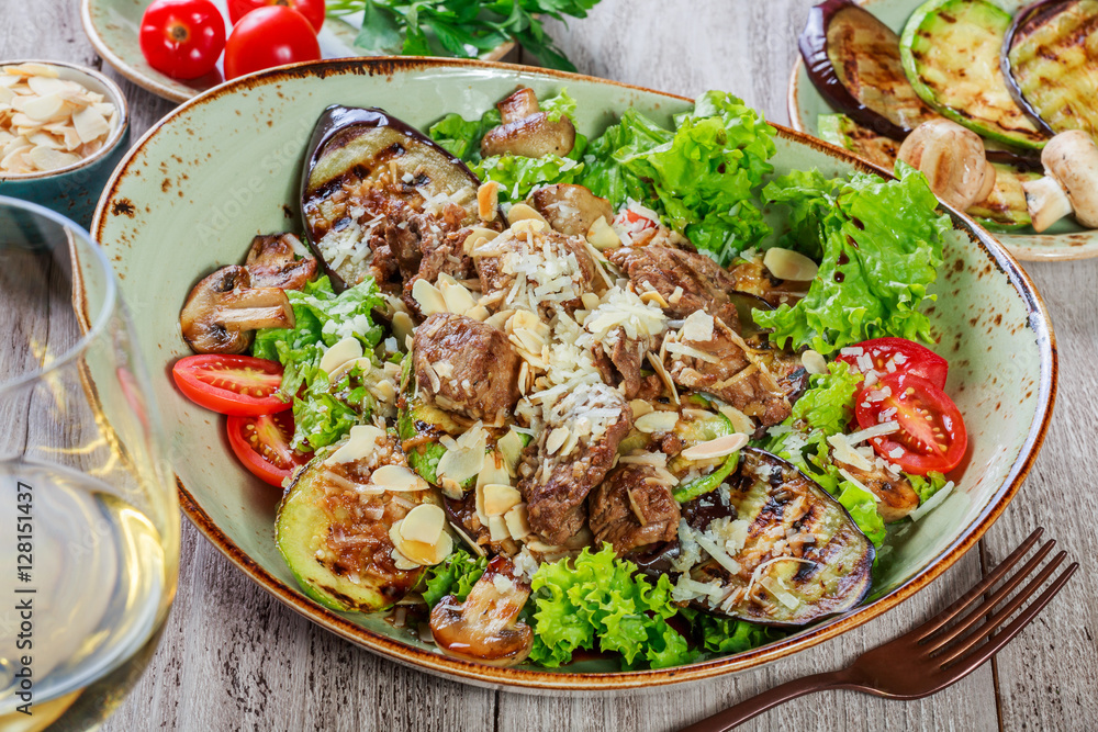 Hot salad with veal, mushrooms, salad leaves, eggplant, zucchini, tomatoes, garnished with grated almonds and Parmesan cheese and glass of wine on wooden background. Healthy food Hot salad with veal, mushrooms, salad leaves, eggplant, zucchini, tomatoes, garnished with grated almonds and Parmesan cheese and glass of wine on wooden background. Healthy food