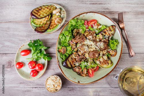 Hot salad with veal, mushrooms, salad leaves, eggplant, zucchini, tomatoes, garnished with grated almonds and Parmesan cheese and glass of wine on wooden background. Healthy food