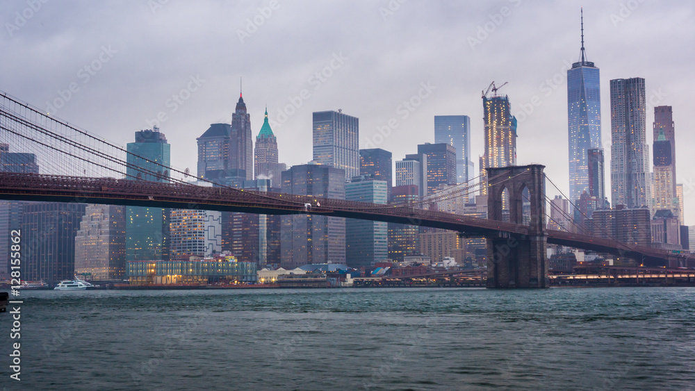Timelapse day to night. Rainy Manhattan and the Brooklyn Bridge. The tops of the skyscrapers in clouds drown. Night comes to the business district of New York City