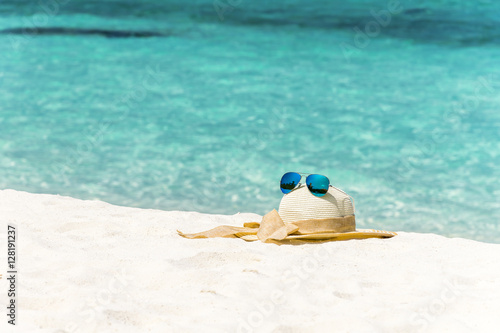 Fototapeta Naklejka Na Ścianę i Meble -  White female trendy hat with blue aviator sunglasses lying on white sand beach in front of turquoise ocean. 