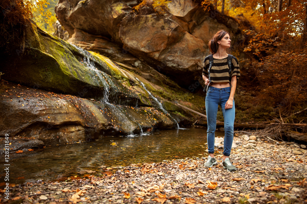 Portrait of a beautiful young woman standing n the forest with a waterfall in the background. Outdoor shot of attractive woman with backpack standing in a mountain stream. Female hiker in creek water.