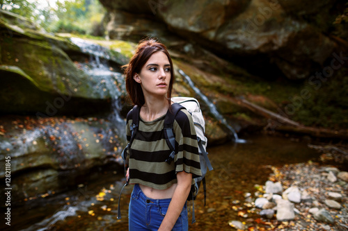 Portrait of a beautiful young woman standing n the forest with a waterfall in the background. Outdoor shot of attractive woman with backpack standing in a mountain stream. Female hiker in creek water.