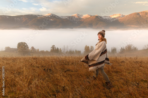 Young woman hiking at mountain peak above clouds and fog Hiker girl wrapping in warm poncho outdoor. Misty mountain. Young girl over the clouds in the valley looking at calm autumn sunrise