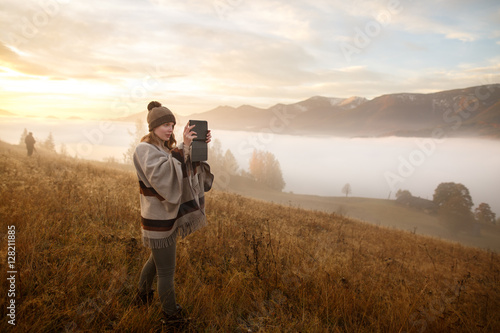 Shot of a young woman looking at the landscape while hiking in the mountains. Hiker taking photo with tablet at mountain peak. Woman in boho style.