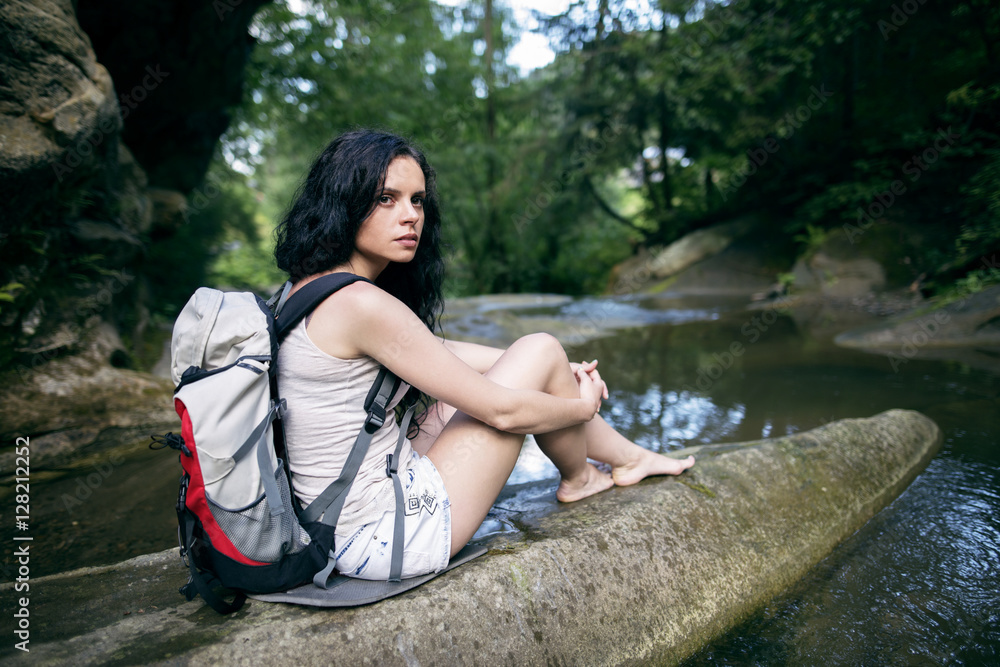 Fototapeta premium Portrait of beautiful young woman with backpack sitting on the rock. Female hiker hiking by the mountain stream.