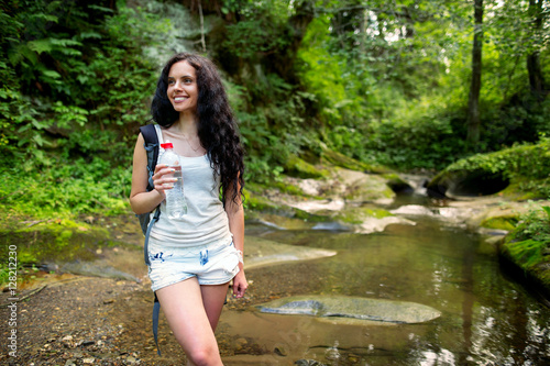 Outdoor shot of attractive young woman with backpack standing in a mountain stream. Female hiker in creek water.Portrait of young pretty brunette woman holding bottle of water.
