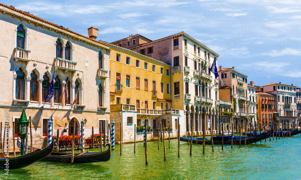 Obraz premium Venice canal with green water and blue sky, colorful houses yellow orange with poles and gondolas and flowers