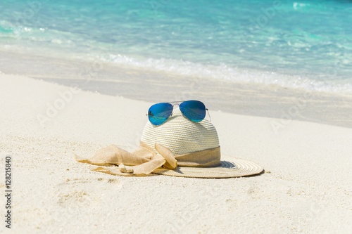 White female trendy hat with blue aviator sunglasses lying on white sand beach in front of turquoise ocean.