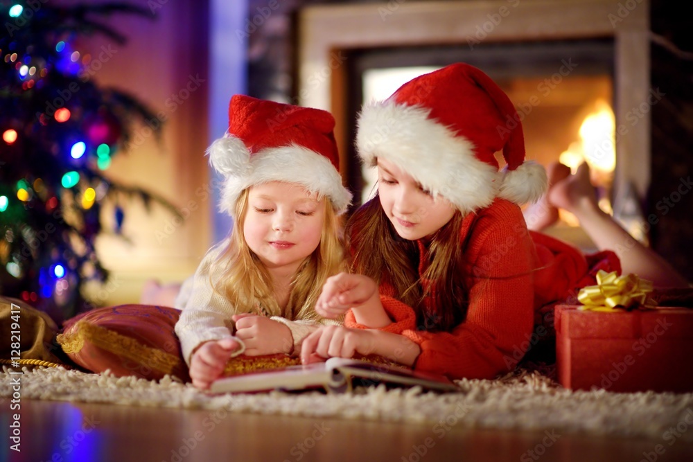 Two cute little sisters reading a story book together under a Christmas tree Two cute little sisters reading a story book together under a Christmas tree