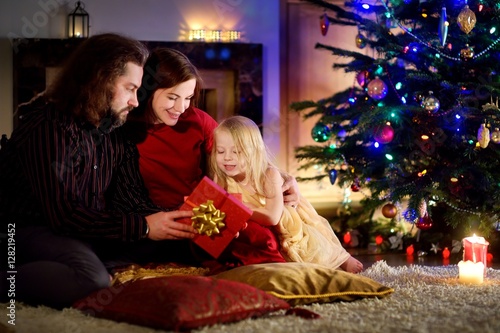 Young happy family of three unwrapping Christmas gifts by a fireplace