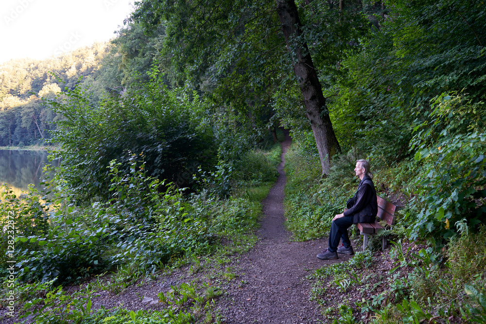 Fotografie Man sitting on a bench beside footpath in the forest near the river