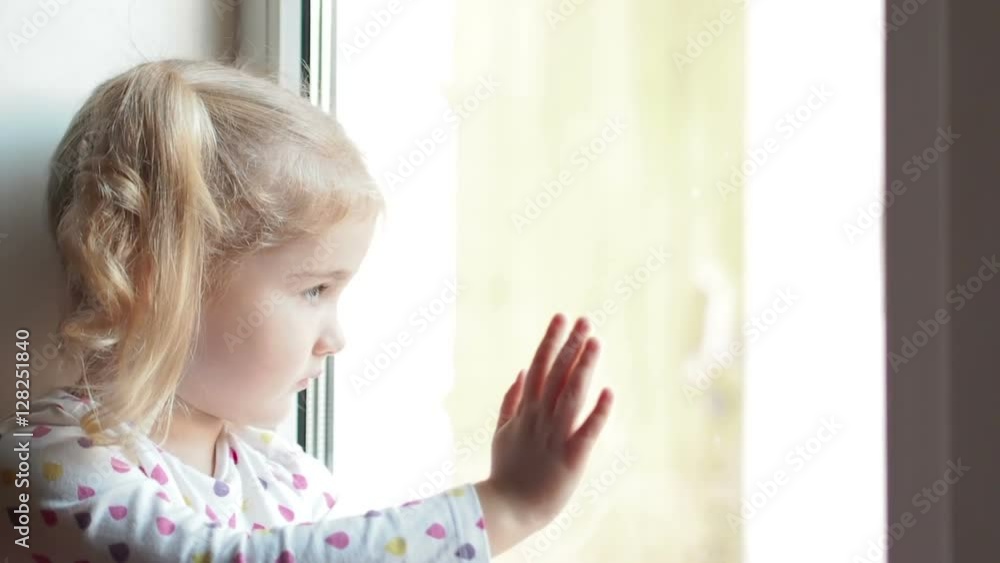 Little girl stares sadly out of a window. Sitting on the window sill. The child looks out of the window. Little girl stares sadly out of a window. Sitting on the window sill. The child looks out of the window.