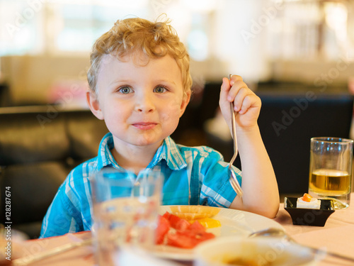 Little blond kid in plaid blue shirt is having fruit breakfast in the resort cafe