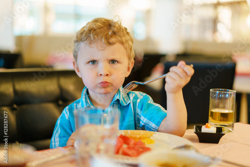 Blond kid is trying taste of fruits with suspicious wary expression on his face. Holding fork in the hand. Dressed in a blue shirt. Fruit bowl and glass of juice on a table.