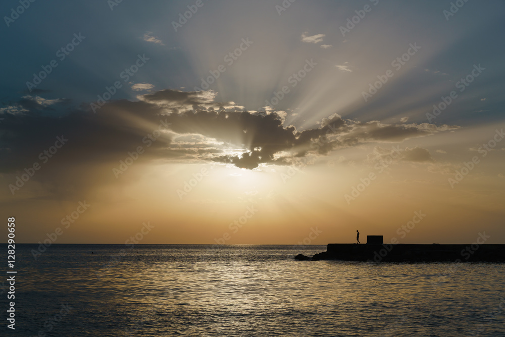 Rays of the evening sun over the calm sea, rapidly breaking through the clouds. Lonely silhouette of a man standing on the breakwater in the distance.