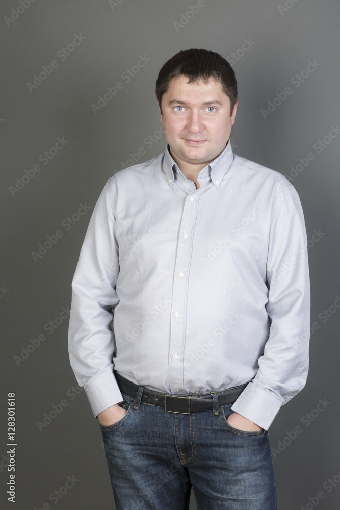 Portrait of a man is in front of the gray background. The man wear jeans and the shirt. 