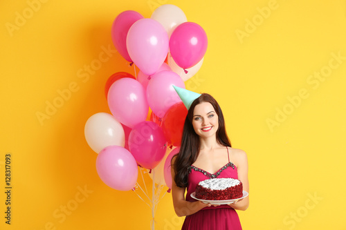 Beautiful young woman in party cap with tasty cake and air balloons on yellow background
